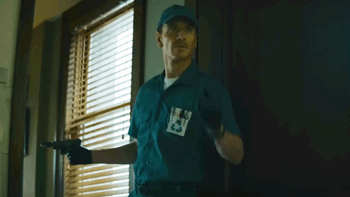 A Man In A Blue Uniform Holds A Gun In Front Of A Window With Blinds .. GIF