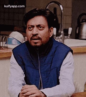 A Man In A Blue Vest Is Sitting At A Table With His Hands Folded In Front Of A Sink In A Kitchen . GIF