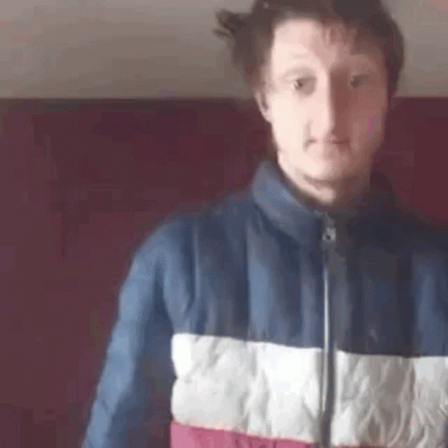 A Man In A Blue , White , And Red Jacket Is Standing In Front Of A Red Wall . GIF