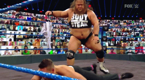 A Man In A Body Of A Steak Shirt Is Standing Over Another Man In A Wrestling Ring GIF