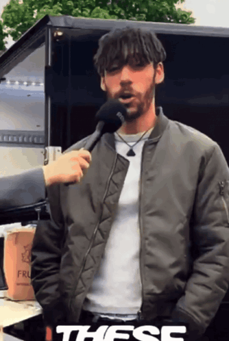 A Man In A Bomber Jacket Is Being Interviewed With A Microphone And The Word These Is On The Bottom Right GIF