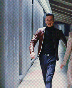 A Man In A Brown Jacket And Black Turtleneck Is Walking Down A Hallway GIF