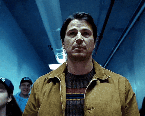 A Man In A Brown Jacket And Sweater Stands In A Hallway GIF