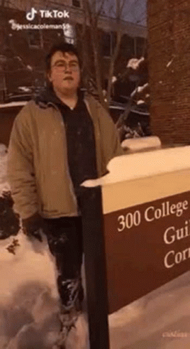 A Man In A Brown Jacket Is Standing Next To A Sign That Says `` 300 College Gui 'S Court '' . GIF
