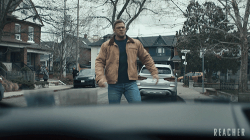 A Man In A Brown Jacket Is Walking Down A Street With The Word Reacher On The Bottom Right GIF