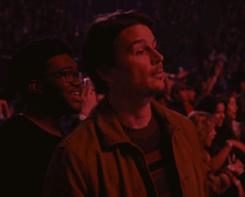 A Man In A Brown Jacket Looks Up At Something In A Crowd GIF