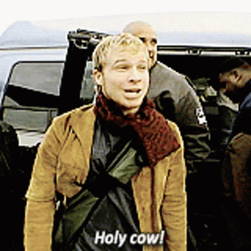 A Man In A Brown Jacket Says Holy Cow In Front Of A Car GIF