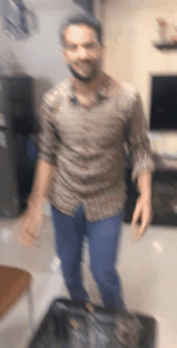 A Man In A Brown Shirt And Blue Jeans Is Standing In A Living Room . GIF