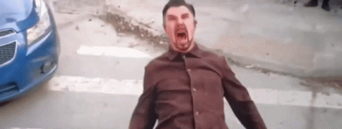 A Man In A Brown Shirt Is Screaming In Front Of A Blue Car On A City Street . GIF