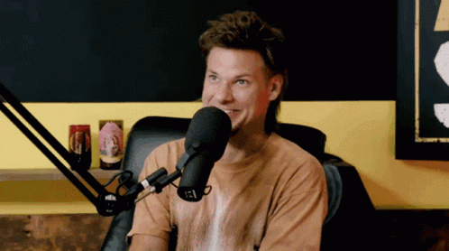 A Man In A Brown Shirt Is Sitting In Front Of A Microphone With A Sticker On It That Says Igl GIF