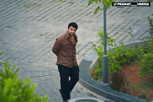 A Man In A Brown Shirt Is Walking Down A Brick Sidewalk With A Sign That Says Pranay_varma GIF