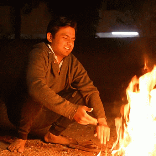 A Man In A Brown Sweater Is Kneeling Down Next To A Fire GIF