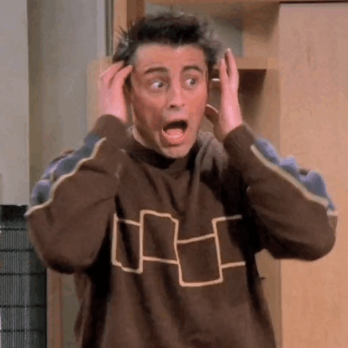 A Man In A Brown Sweater Is Making A Surprised Face While Holding His Hands To His Head . GIF