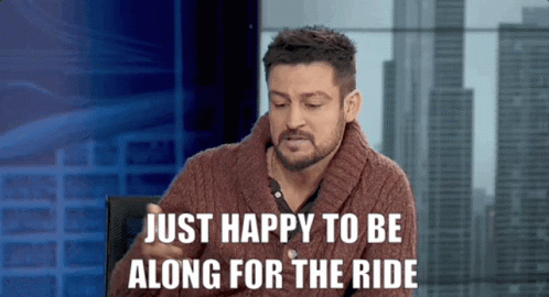 A Man In A Brown Sweater Is Sitting In Front Of A Sign That Says Just Happy To Be Along For The Ride GIF