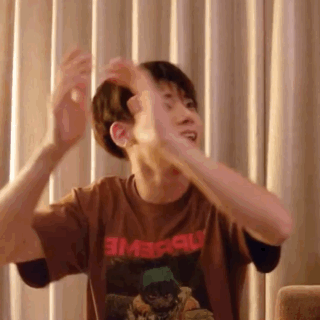 A Man In A Brown T-Shirt Is Sitting On A Couch With His Arms Outstretched In Front Of Curtains . GIF