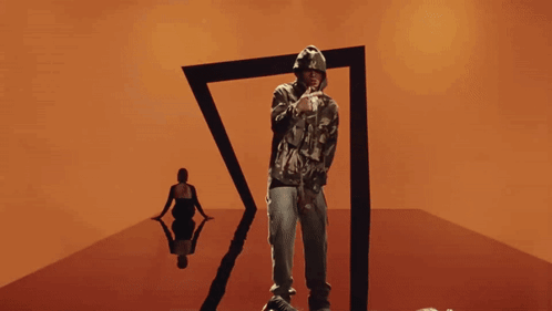 A Man In A Camo Hoodie Stands In Front Of A Black Triangle GIF