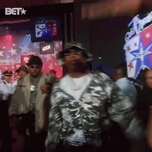 A Man In A Camo Jacket Is Dancing In Front Of A Crowd Of People . GIF