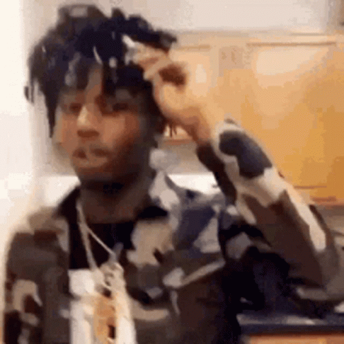 A Man In A Camouflage Jacket Is Standing In A Kitchen With His Hand On His Hair . GIF