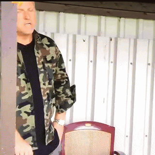 A Man In A Camouflage Shirt Is Standing In Front Of A Chair GIF