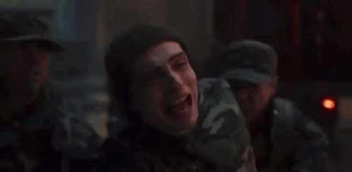 A Man In A Camouflage Uniform Is Laughing In A Dark Room With Other Men . GIF