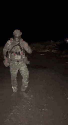 A Man In A Camouflage Uniform Is Standing In The Dark Holding A Gun . GIF
