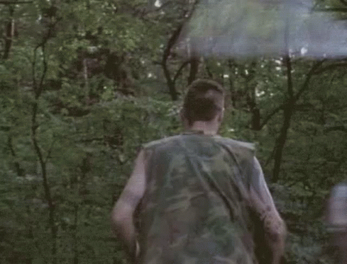 A Man In A Camouflage Vest Is Running Through The Woods With A Smoke Bomb . GIF