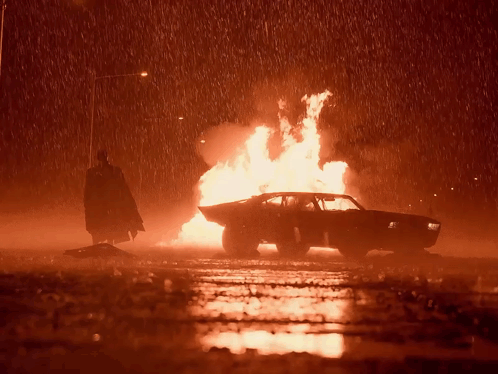 A Man In A Cape Is Standing Next To A Burning Car GIF