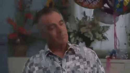 A Man In A Checkered Shirt Is Sitting In Front Of Balloons And Flowers In A Room . GIF