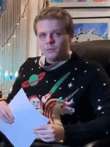 A Man In A Christmas Sweater Is Sitting In A Chair Holding A Piece Of Paper GIF
