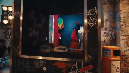 A Man In A Clown Costume Is Standing In A Box With Graffiti On The Wall . GIF
