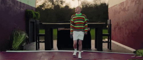A Man In A Colorful Sweater And White Shorts Is Standing In Front Of A Table . GIF