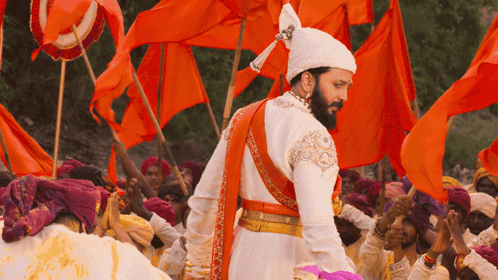 A Man In A Costume With A Beard Stands In Front Of A Crowd Of People Holding Orange Flags GIF