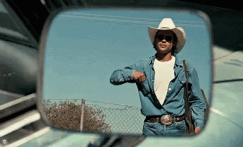 A Man In A Cowboy Hat And Sunglasses Is Standing In Front Of A Car In A Rear View Mirror . GIF