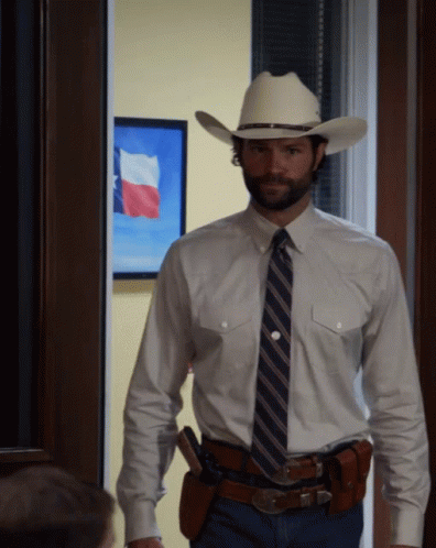 A Man In A Cowboy Hat And Tie Stands In Front Of A Painting Of A Texas Flag GIF