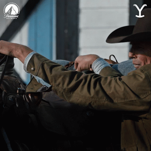 A Man In A Cowboy Hat Is Holding A Blanket In Front Of A Paramount Sign GIF