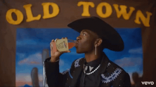 A Man In A Cowboy Hat Is Holding A Drink In Front Of An Old Town Poster GIF