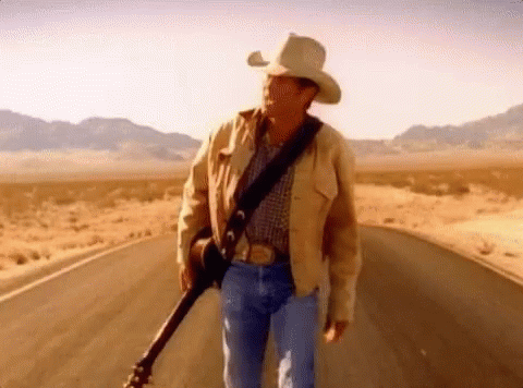 A Man In A Cowboy Hat Is Holding A Guitar While Walking Down A Road . GIF