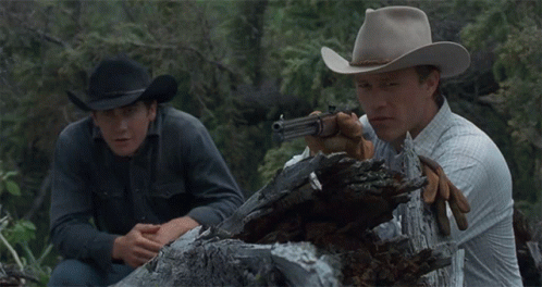 A Man In A Cowboy Hat Is Holding A Gun Next To Another Man GIF