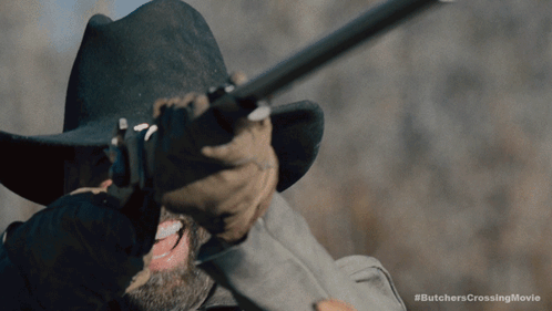 A Man In A Cowboy Hat Is Holding A Rifle With The Hashtag #Butcherscrossingmovie Below Him GIF
