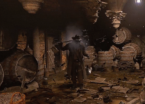 A Man In A Cowboy Hat Is Standing In A Dark Room With Barrels GIF