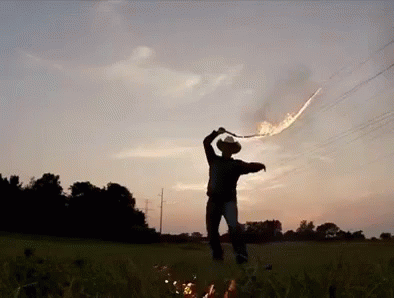 A Man In A Cowboy Hat Is Standing In A Field Holding A Sword With Flames Coming Out Of It GIF