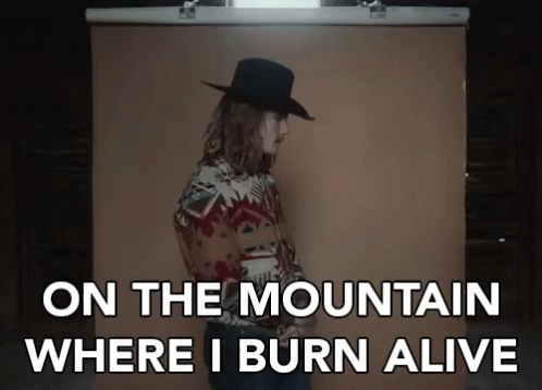 A Man In A Cowboy Hat Is Standing In Front Of A Brown Backdrop With The Words On The Mountain Where I Burn Alive GIF