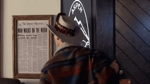 A Man In A Cowboy Hat Is Standing In Front Of A Framed Newspaper That Says Man Walks On The Moon GIF