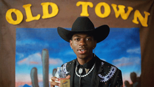 A Man In A Cowboy Hat Is Standing In Front Of An Old Town Sign GIF