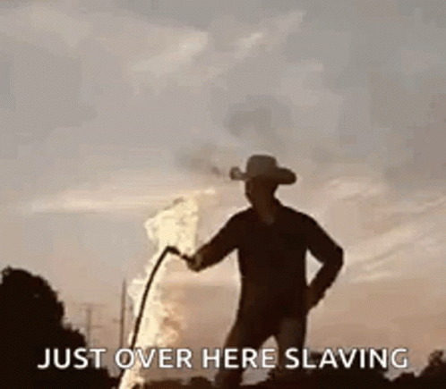 A Man In A Cowboy Hat Is Standing Next To A Fire With A Hose . GIF