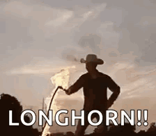 A Man In A Cowboy Hat Is Standing Next To A Fire With The Words `` Longhorn '' Written On It . GIF
