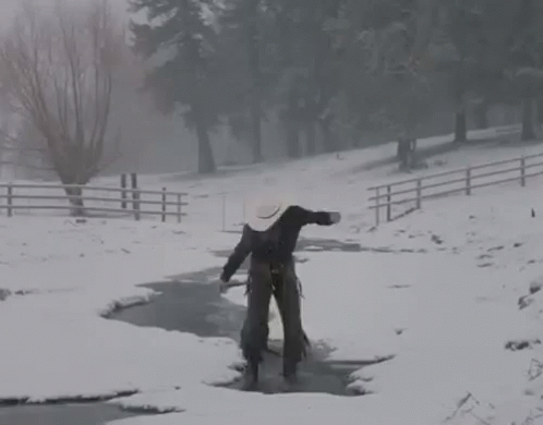 A Man In A Cowboy Hat Is Walking Across A Snow Covered Stream . GIF