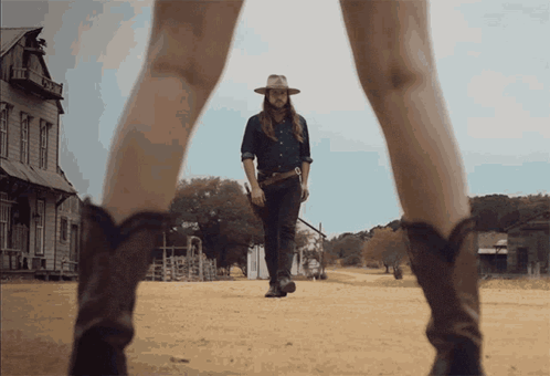 A Man In A Cowboy Hat Is Walking Down A Dirt Road Next To A Woman In Cowboy Boots GIF