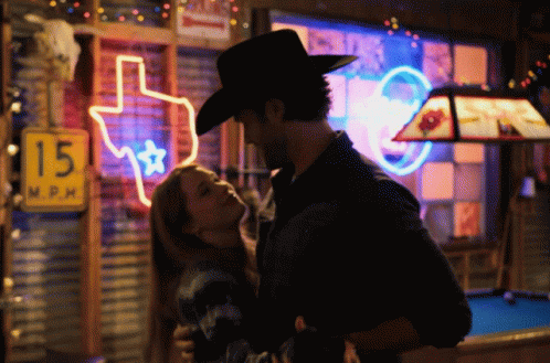A Man In A Cowboy Hat Kisses A Woman In Front Of A Neon Sign That Says 15 Mph GIF