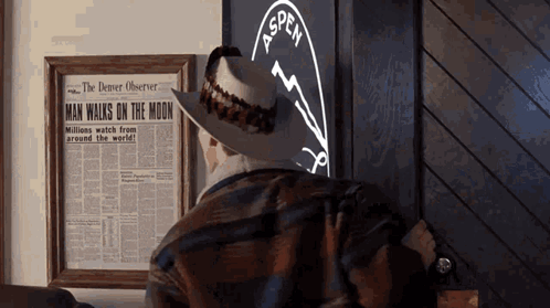 A Man In A Cowboy Hat Looks At A Framed Newspaper That Says Man Walks On The Moon GIF